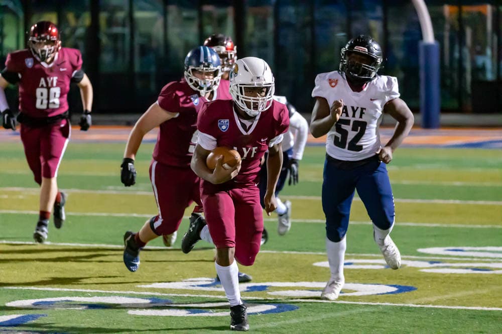 AYF football players running during a night game, the ball carrier in a maroon jersey